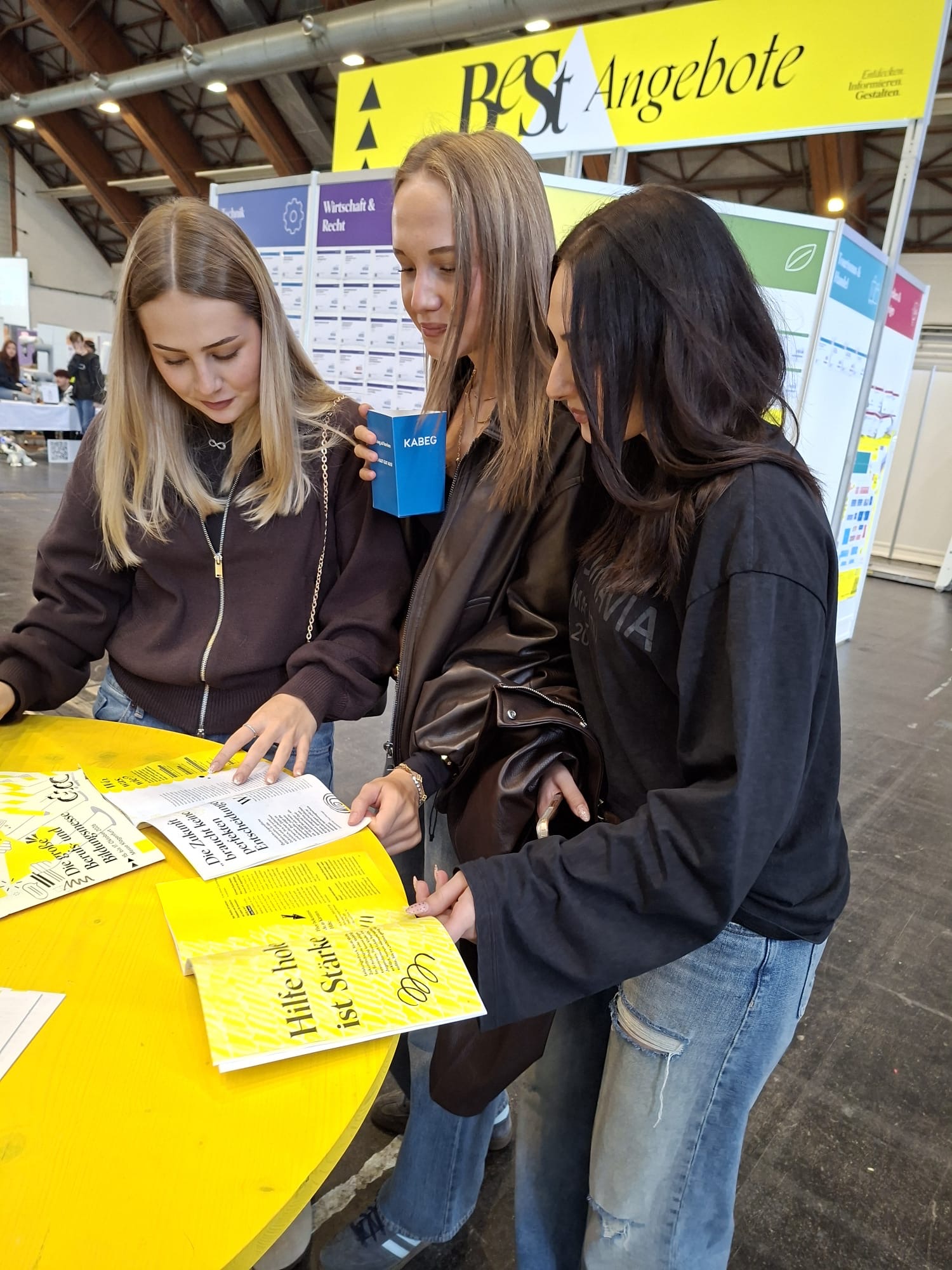 Drei junge Frauen lesen gelbe Broschüren an einem runden Tisch.